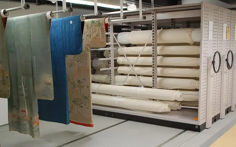 Traditional garments hang on a rack to the left, while neatly rolled fabrics are stored on shelves to the right. The setting appears to be a textile storage area or museum collection.