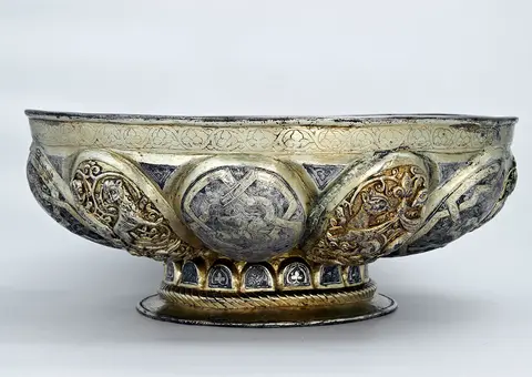 A decorative bowl featuring intricate silver and gold designs. The bowl is embellished with ornate patterns and reliefs, displaying mythical figures and foliage motifs. It has a wide, shallow shape with a raised base, showcasing skilled craftsmanship.