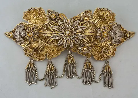 Intricately designed vintage brooch featuring a floral centerpiece surrounded by ornate textures in gold and silver tones. The piece is adorned with small gemstones and hangs with several silver tassels, showcasing detailed craftsmanship.