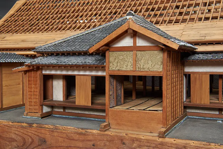 A detailed wooden model of a traditional Japanese house, showcasing intricate architectural elements such as a tiled roof, sliding doors, and wooden beams. The model conveys a sense of historical craftsmanship and design typical of Japanese residential structures.