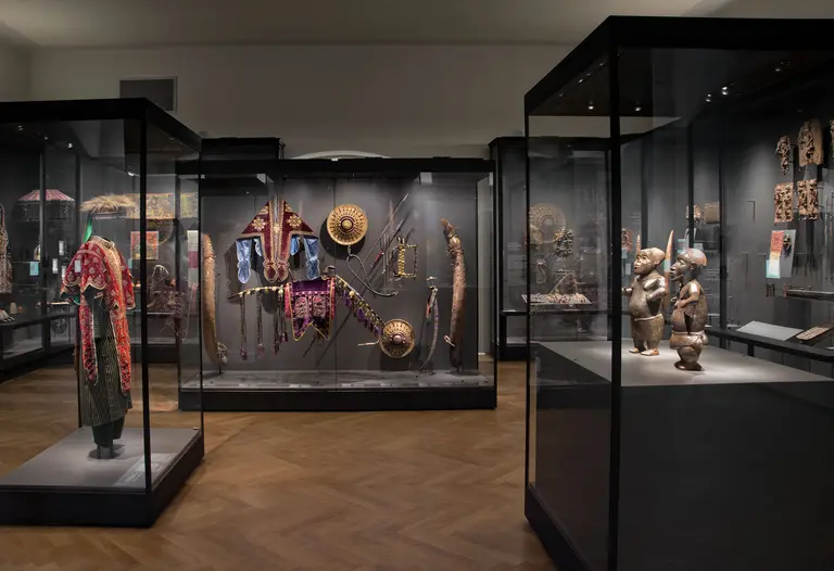 Das Bild zeigt einen Ausstellungsraum mit Vitrinen, die verschiedene kunstvolle Objekte und traditionelle Kleidung präsentieren. Zu sehen sind Masken, Schilde und Figuren, die kulturelle Artefakte darstellen. Der Boden ist aus Holz, und die Wände sind dunkel gehalten, um die Exponate hervorzuheben.