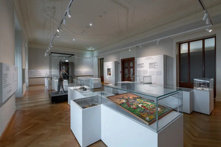 Ein Ausstellungsraum mit mehreren Vitrinen, die Kunstobjekte zeigen. Im Hintergrund ist ein Besucher zu sehen, der sich die Exponate anschaut. Die Wände sind mit Erklärungen und Informationen zur Ausstellung dekoriert. Helle Beleuchtung und ein eleganter Holzfußboden ergänzen die Atmosphäre.