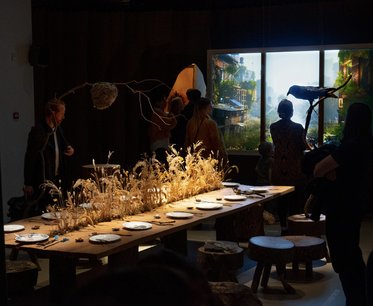 Eine gedeckte Tafel mit braunem Tisch und Tellern, umgeben von Dürren Pflanzen. Menschen stehen und interagieren im Raum, während im Hintergrund ein großes Fenster eine grüne Landschaft zeigt. Die Atmosphäre ist kreativ und einladend.