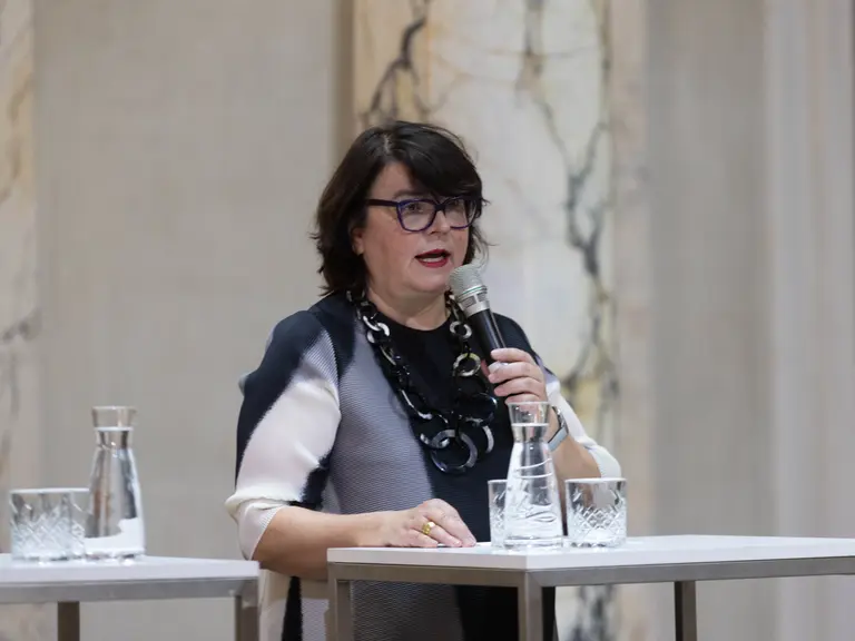 Eine Frau mit Brille spricht in ein Mikrofon, während sie an einem Tisch steht. Auf dem Tisch sind Wassergläser und eine Karaffe platziert. Im Hintergrund sind helle, marmorierte Wände zu sehen.