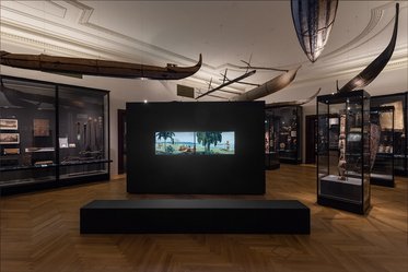 A museum interior featuring various exhibits, including displayed artifacts in glass cases. In the center, a large black panel showcases a landscape image with palm trees and a boat. Above, traditional boats hang from the ceiling, while the floor is wooden.
