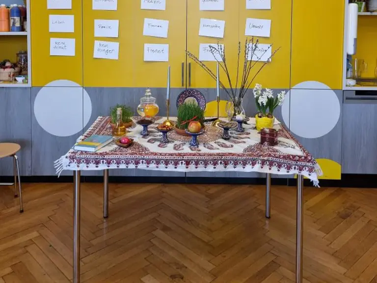 Ein gedeckter Tisch mit traditioneller Tischdecke, dekoriert mit Schalen voller Obst, Gläsern mit Getränken und frischen Kräutern. Im Hintergrund eine gelbe Wand mit Notizen. Die Szene vermittelt eine festliche Atmosphäre, möglicherweise für ein kulturelles Fest oder ein gemeinsames Essen.