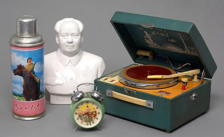 A vintage green phonograph with a red turntable sits open, next to a white porcelain bust of a man. Also displayed are a silver spray can featuring a colorful image and a round alarm clock with a cartoon character. The background is plain gray.