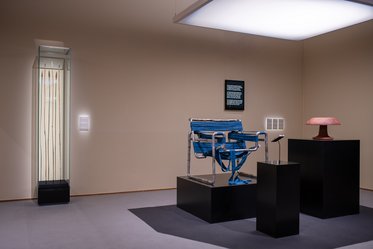Ein modernes Ausstellungszimmer zeigt einen blauen Stuhl aus Metall und Stoff auf einem schwarzen Podest. Neben dem Stuhl steht eine rote Lampe auf einem weiteren Podest. Im Hintergrund ist ein Glasdisplay mit länglichen Objekten und einer Informationstafel zu sehen.