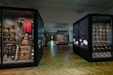 An interior view of a museum showcasing various displays. There are glass cases filled with intricate artifacts, textiles, and sculptures. The wooden floor features a herringbone pattern, and the space is well-lit, highlighting the exhibits.