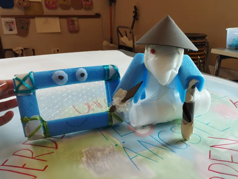 Eine Figur aus Schaumstoff, gekleidet in blau, hält einen Pinsel und trägt einen Hut. Neben ihr steht ein rechteckiger Bilderrahmen aus Schaumstoff mit googly Augen. Der Hintergrund zeigt ein Blatt mit farbigen Schriftzügen.