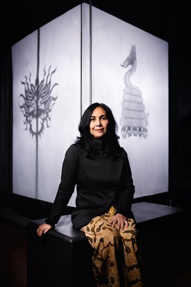 A woman with long black hair sits on a bench, wearing a black top and patterned golden pants. Behind her, a large illuminated cube casts shadows of intricate designs, including a dragon and a sun motif, onto the wall. The setting is dimly lit, emphasizing the shapes.