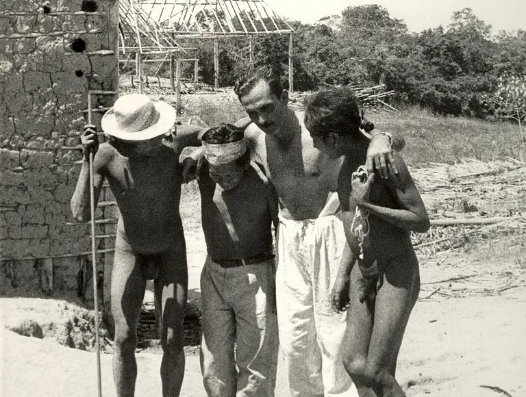 Eine Gruppe von vier Personen, darunter zwei Männer und zwei Frauen, versammelt sich in einer ländlichen Umgebung. Sie stehen eng beieinander und scheinen miteinander zu sprechen oder zu lachen. Im Hintergrund ist ein roh errichtetes Gebäude sichtbar.
