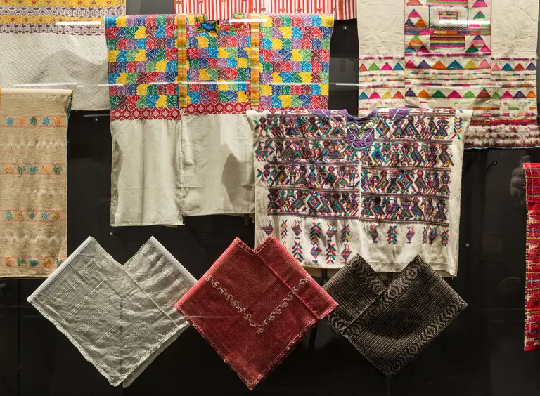 Eine Sammlung traditioneller Textilien ist zu sehen, bestehend aus bunten, gemusterten und einfarbigen Stoffen. Die Stoffe weisen verschiedene Muster und Farben auf, die kulturelle Motive darstellen, und sind an einer Wand aufgehängt.