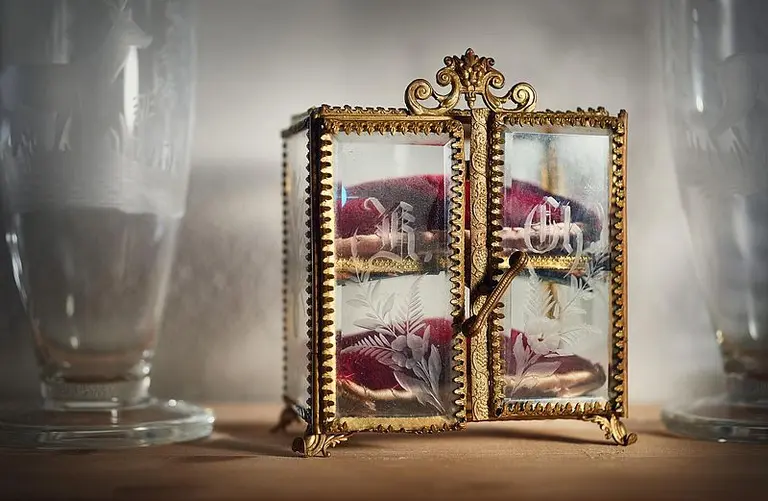 Ein kleiner, eleganter Glasschrank mit goldenen Verzierungen steht auf einem Tisch. Die Türen sind teilweise geöffnet und zeigen kunstvoll gestaltete Innenteile mit bunten Stoffen und floralen Mustern. Zwei große transparente Gläser flankieren den Schrank.