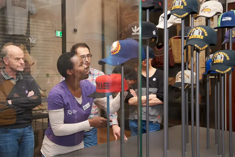 Eine Museumführerin erklärt eine Gruppe von Besuchern verschiedene Baseballkappen, die in einem Ausstellungsglas präsentiert werden. Die Besucher hören aufmerksam zu und beobachten die Kappen, während sie sich um die Führerin versammeln.