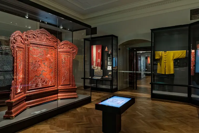 Ein Museumsaal mit Vitrinen. Im Vordergrund steht ein kunstvoll geschnitzter, roter Holzschrank. Neben ihm befinden sich ein interaktives Display und weitere ausgestellte Objekte, darunter ein gelbes Gewand, das an der Wand hängt. Der Boden ist aus Holz, und die Wände sind neutral gehalten.