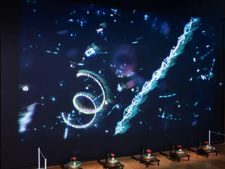 Eine große Projektion zeigt abstrakte, leuchtende Muster und Formen auf einer Wand. Im Vordergrund sind mehrere Geräte platziert, die auf Tischplatten stehen. Die Atmosphäre wirkt futuristisch und künstlerisch, mit Farben, die in der Projektion lebendig strahlen.