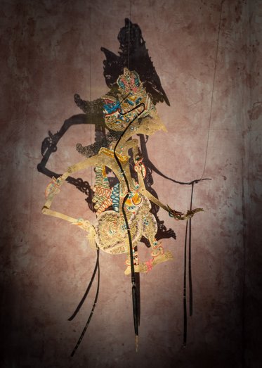 A colorful shadow puppet casting a detailed silhouette on a textured brown background. The puppet features intricate designs and is depicted in an upright position, suggesting movement or performance.