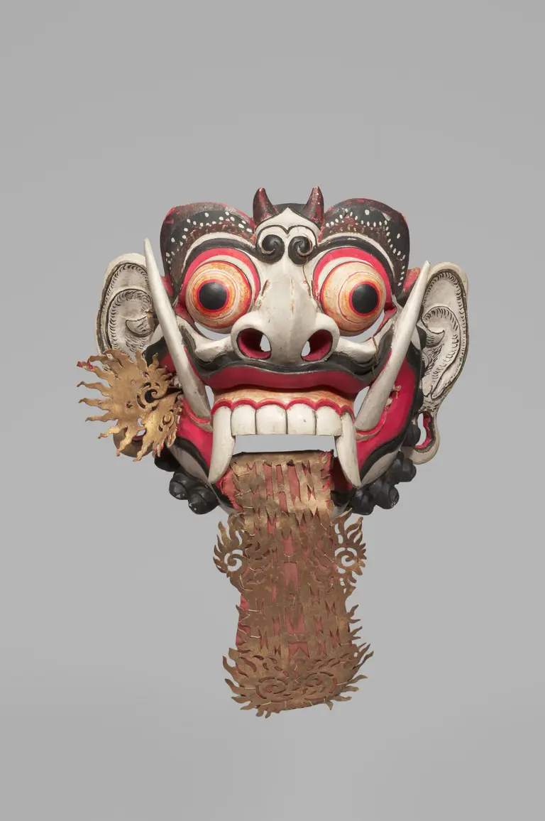 Ein bunter, kunstvoll gestalteter Maskenkopf mit großen, runden Augen, spitzen Zähnen und auffälligen Ohren. Die Maske zeigt eine Kombination aus roten, weißen und schwarzen Farben sowie ornamentalen Verzierungen. Von der Unterseite hängt ein dekorierter Streifen aus geschnitztem Material herab.