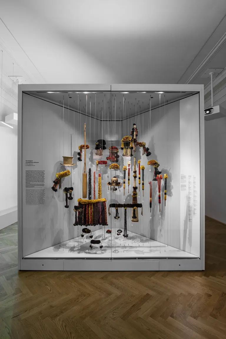 Eine Glasvitrine zeigt eine künstlerische Installation mit verschiedenen Objekten, die von oben hängen. Die Objekte bestehen aus leuchtenden Materialien in unterschiedlichen Farben und Formen. Die Vitrine befindet sich in einem Ausstellungsraum mit Holzfußboden und neutralen Wänden.