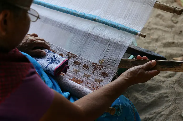 Eine Person sitzt an einem Webstuhl und arbeitet an einer Webarbeit. Sie hält einen Holzstab in der Hand und konzentriert sich auf das gewebte Muster, das Tiere darstellt. Die Umgebung ist einfach, mit einem bunten Stoff in ihrer Nähe.