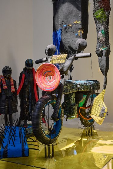 Eine künstlerische Installation zeigt ein Fahrrad aus recycelten Materialien, umgeben von stilisierten Figuren in auffälligen Kleidern. Verschiedene Texturen und Farben dominieren die Szene, mit einem Fokus auf Nachhaltigkeit und Kreativität in der Kunst.
