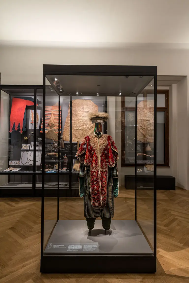Eine kostbare historische Tracht in einem Glasvitrinen-Ausstellungsstück. Die Kleidung ist reich verziert, mit roten und goldenen Elementen, und wird von einem Hut ergänzt. Im Hintergrund sind weitere Exponate und Schriftstücke sichtbar, die zur Ausstellung gehören.