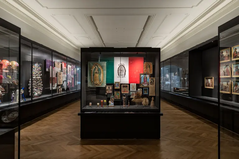 Das Bild zeigt eine Ausstellungshalle mit Vitrinen, in denen verschiedene kulturelle Artefakte aus Mexiko präsentiert werden. Im Zentrum ist eine Vitrine mit einer mexikanischen Flagge und religiösen Bildern, umgeben von weiteren Exponaten entlang der Wände. Der Boden ist aus Holzparkett.