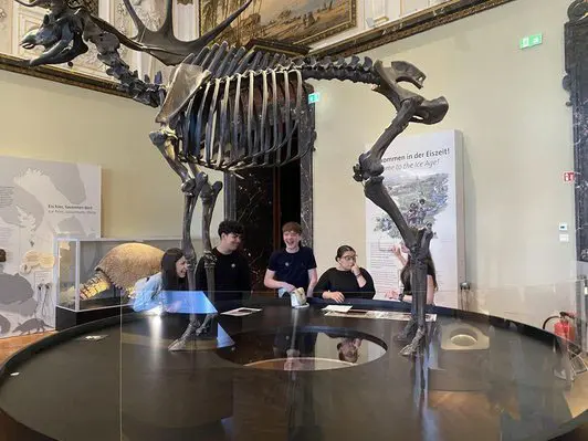 Das Bild zeigt eine Gruppe von fünf Personen in einem Museum, die an einem runden Tisch stehen und sich mit einem Skelett eines Dinosauriers im Hintergrund beschäftigen. Das Skelett ist eindrucksvoll und dominiert den Raum. Informationen zu den Exponaten sind an der Wand sichtbar.
