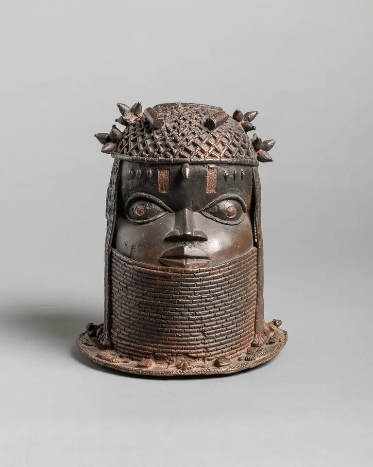 Eine bronzene Skulptur des Kopfes, verziert mit einem geflochtenen Kopfschmuck und stacheligen Elementen. Die durchdringenden Augen und der breite Mund verleihen der Figur eine ausdrucksstarke Präsenz. Die Details der Textur sind gut ausgearbeitet und die Form ist stark und markant.