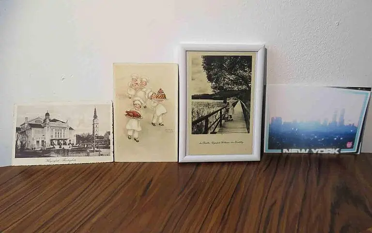 Auf dem Bild sind vier Bilderrahmen nebeneinander angeordnet. Dazu gehören eine historische Ansichtskarte eines Gebäudes, eine Zeichnung von zwei Mädchen mit Bonbons, ein gerahmtes Foto einer Holzbrücke in der Natur und eine moderne Darstellung von New York City.