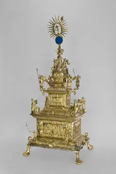 A detailed golden ornate reliquary featuring intricate sculptures of angels and figures, topped with a sunburst motif and a blue circular centerpiece. It stands on four feet, showcasing elaborate decorative elements and embellishments throughout its structure. The background is a plain gray, emphasizing the piece's craftsmanship.