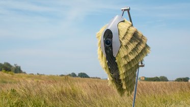 Eine moderne Maschine mit einem gefiederten Design steht auf einem Ständer in einem Feld. Im Hintergrund erstreckt sich eine weitläufige Landschaft mit Grün- und Brauntönen unter einem blauen Himmel.