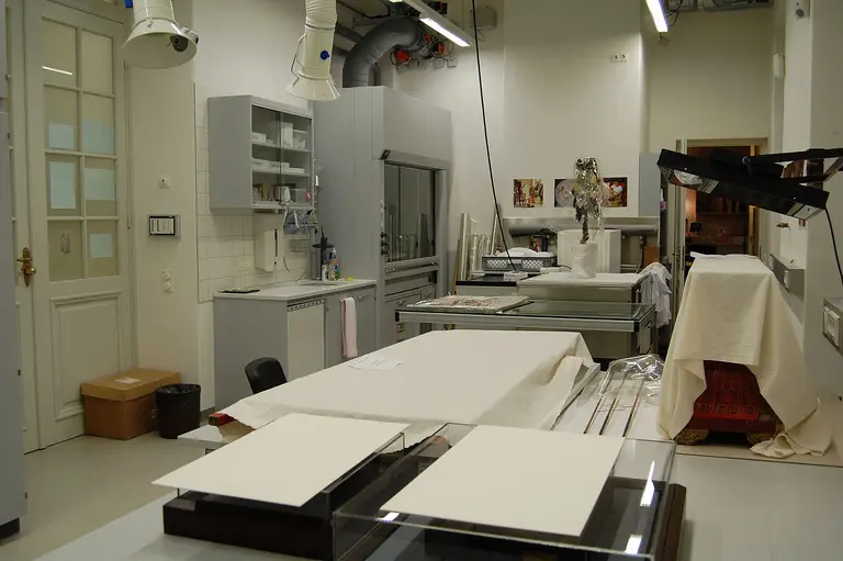 A spacious laboratory with countertops and equipment for preservation work. The room features shelves with various supplies, a large worktable, and several lit workstations. White sheets cover some surfaces, and there are tools and materials visible, hinting at ongoing restoration activities.