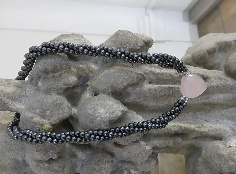 Eine Halskette mit schwarzen Perlen und einem rosa Anhänger, die elegant auf einer steinernen Oberfläche drapiert ist. Die Kette zeigt einen Kontrast zwischen den dunklen Perlen und dem sanften Glanz des Anhängers.