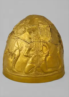 A gold decorative helmet featuring intricate reliefs. The artwork depicts two armored figures, one kneeling and the other holding a weapon, set against a curved background. The helmet has a smooth surface with detailed engravings, showcasing historical craftsmanship.