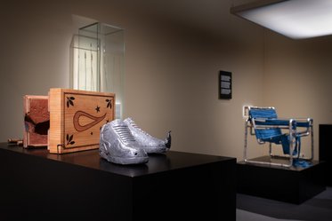 Eine silberne Sportschuhskulptur liegt auf einem schwarzen Podest. Neben den Schuhen befindet sich eine holzverzierte Kiste. Im Hintergrund steht ein moderner, blauer Stuhl und eine Informationstafel an der Wand.