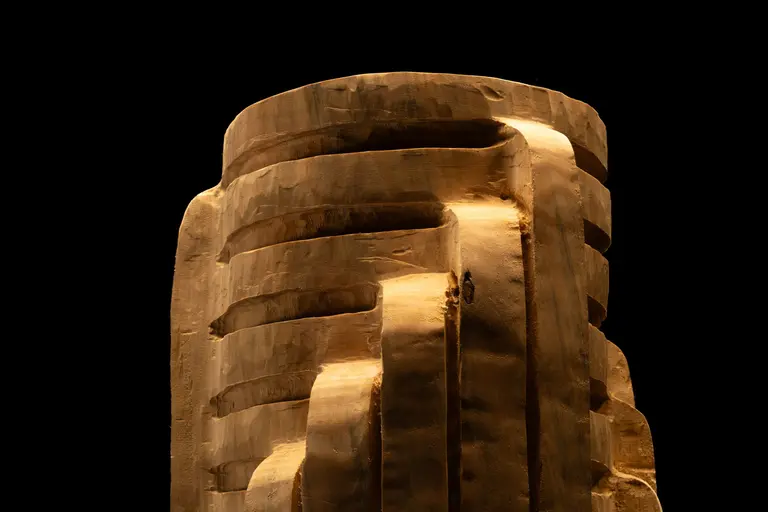 A large, textured sculpture resembling a tall, abstract structure with curved lines and steps. The sculpture is illuminated against a black background, emphasizing its intricate details and shadows. A small figure can be seen on the sculpture, providing a sense of scale.