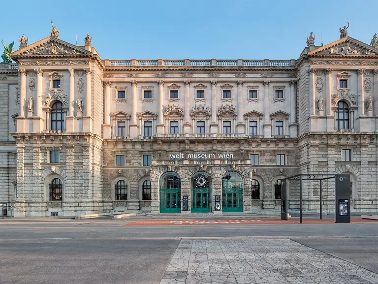 Das Bild zeigt die beeindruckende Fassade des Weltmuseums Wien. Das historische Gebäude hat eine aufwändige Architektur mit Statuen und Säulen, sowie großen Eingängen. Am Gebäude ist der Schriftzug 