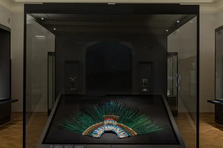 Ein prächtiger Federkopfschmuck ist in einer Vitrine ausgestellt. Er besteht aus bunten Federn, die in einem bogenförmigen Muster angeordnet sind. Die Vitrine befindet sich in einem dunklen Raum, wobei im Hintergrund weitere Ausstellungsobjekte sichtbar sind.