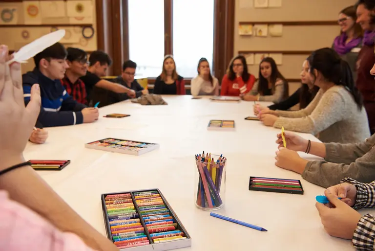 Eine Gruppe von Jugendlichen sitzt an einem Tisch mit Kunstmaterialien, darunter Buntstifte und Kreiden. Sie sind in angeregter Unterhaltung vertieft, während einige mit Farbstiften arbeiten. Der Raum hat große Fenster, die Tageslicht hereinlassen.