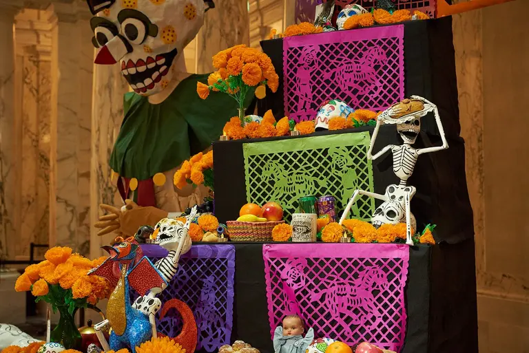 Ein Altarschrein für den Día de los Muertos zeigt leuchtend orangefarbene Marigoldblüten, dekorierte Skelette und traditionelles Obst. Bunte Stoffe und ein großes Maskottchen im Hintergrund verleihen dem Arrangement Feierlichkeit und kulturelle Bedeutung. Der Altar trägt Bilder und Gegenstände zur Ehrung der Verstorbenen.