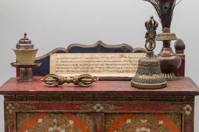 Auf einem dekorativen Tisch stehen verschiedene religiöse Artefakte: eine goldene Glocke, eine Skulptur mit bunten Federn, ein Schriftstück mit fremden Schriftzeichen und ein zeremonieller Stab. Die Gegenstände sind kunstvoll verziert und spiegeln kulturelle Traditionen wider.