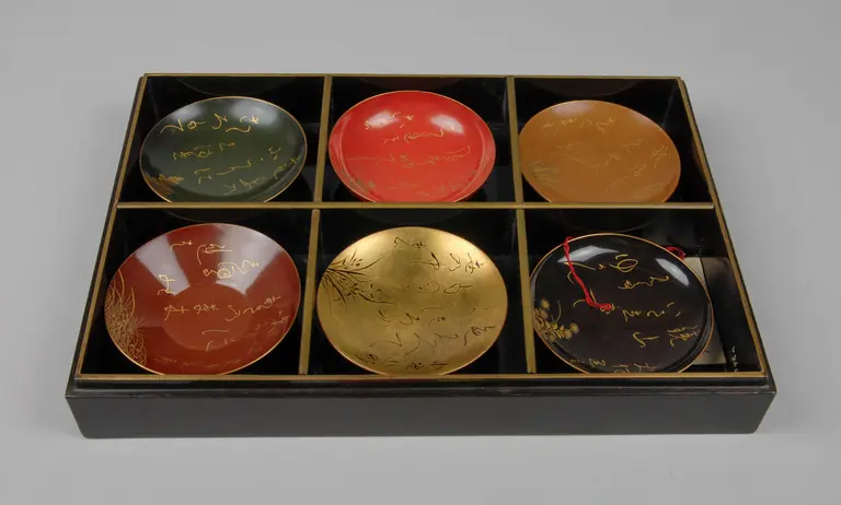 A black box contains six decorative dish plates, each with intricate gold or colored designs. The plates feature varying colors, including red, gold, and dark hues, with some displaying elegant inscriptions. The arrangement suggests a curated collection of artistic craftsmanship.