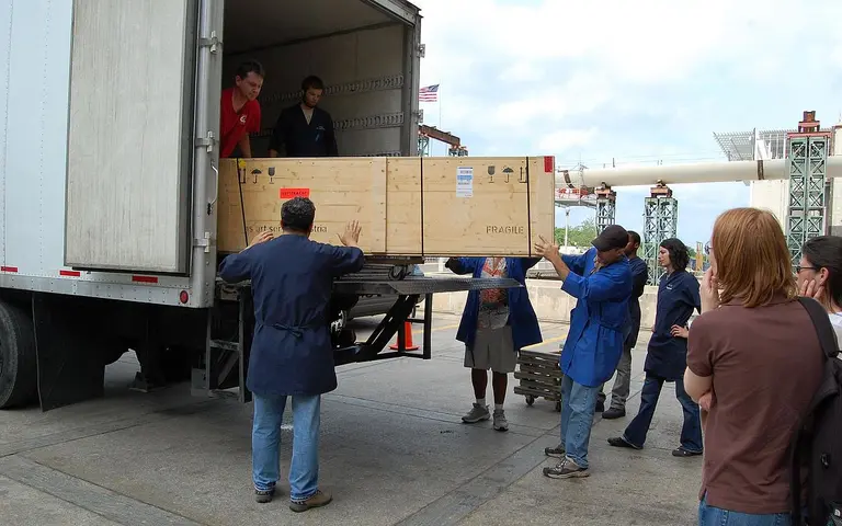 A group of six people is unloading a large wooden crate from the back of a truck. Some individuals are holding the crate while others assist in the process. The setting appears to be an outdoor loading area, with a cloudy sky above.
