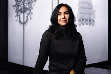 A woman with long black hair sits confidently, wearing a black outfit. She is positioned in front of softly illuminated backgrounds displaying intricate shadow patterns, contributing to a warm and artistic ambiance.