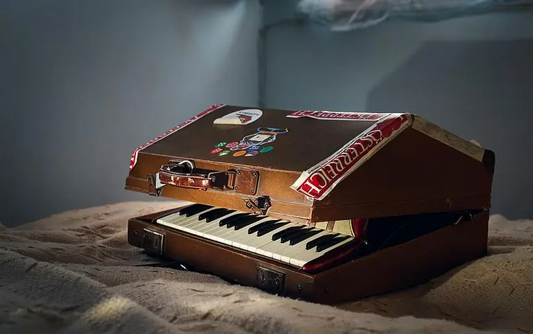 Ein geöffnetes Klavier im Kofferformat mit schwarzen und weißen Tasten. Der Koffer ist mit verschiedenen Aufklebern dekoriert und liegt auf einer weichen Unterlage. Im Hintergrund ist ein schwaches Licht zu sehen.