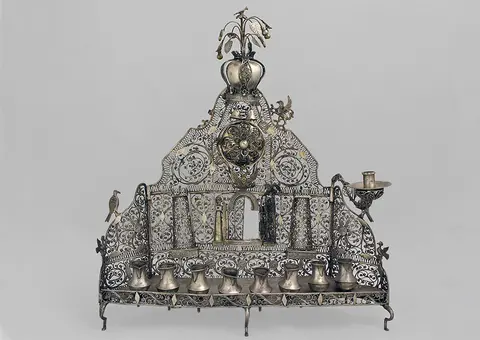 A decorative silver menorah with intricate filigree design. It features a central circular element, ornate floral and bird motifs, and eight candle holders along the bottom, a larger holder on the right for the shamash candle. The background is plain for contrast.