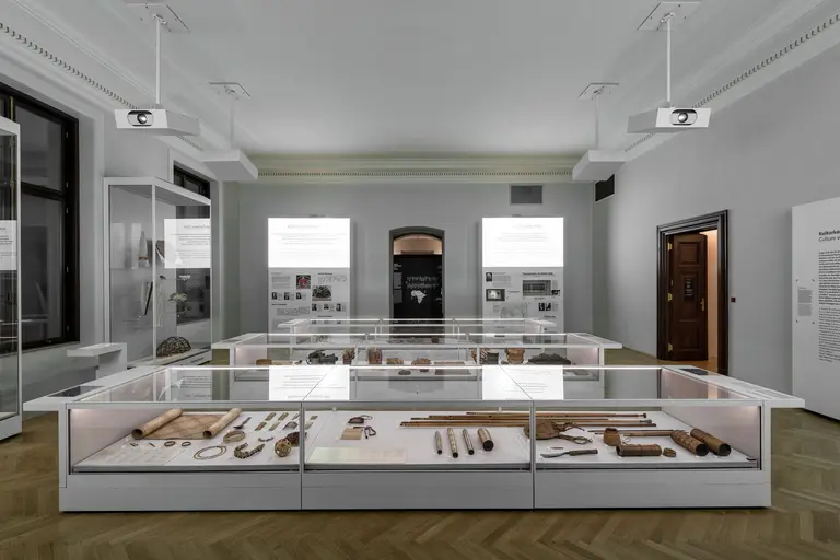 In einem Museumsaal sind Ausstellungsstücke in vitrinen angezeigt. Die Vitrinen enthalten verschiedene historische Objekte, während Informationstafeln an der Wand beschrieben sind. Der Raum ist hell erleuchtet und wirkt einladend.