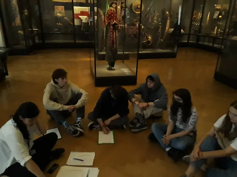 Sechs Personen sitzen im Kreis auf dem Boden eines Museums. In der Mitte liegen Notizen und ein grünes Heft. Im Hintergrund ist ein Vitrinenschrank zu sehen, der ein historisches Outfit oder eine Figurenstatue zeigt. Das Licht ist gedämpft und die Atmosphäre ist ruhig.